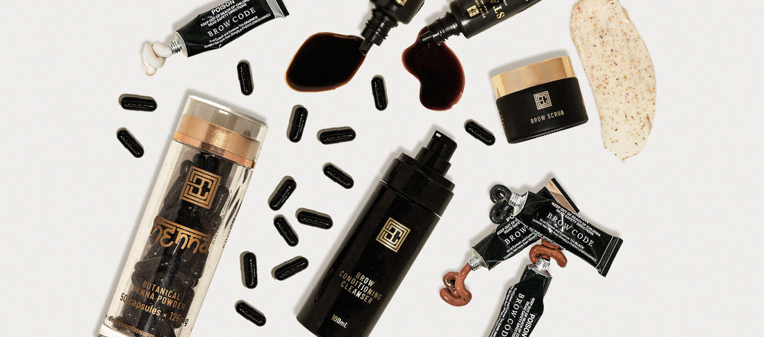 The image shows a flat lay of Brow Code products, including Henna Powder, Brow Conditioner Cleanser, Brow Tint tubes and Brow Stain liquid spills and a Brow Scrub smear on a light background.